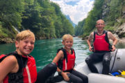 Smiling, happy father and two sons sitting in a grey raft on the Tara river on a partly cloudy partly sunny day in Montenegro, with a waterfall behind them