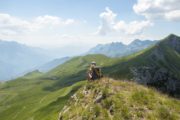 A solo female hiker sitting on a remote and grassy mountain peak in Montenegro, looking out at a stunning view