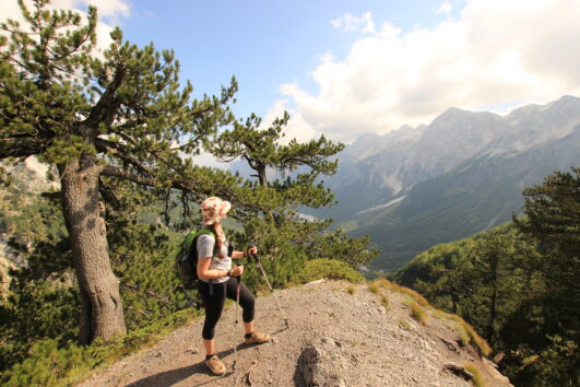 A solo hiker looks out, on a part sunny, cloudy day, onto Valbona valley below, on the pass from Theth, part of the Albania section of the Peaks of the Balkans hiking trail.