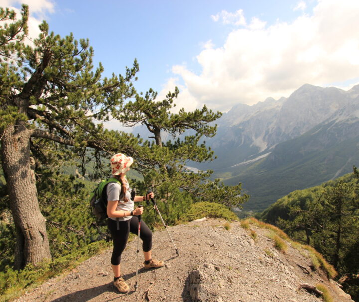 A solo hiker looks out, on a part sunny, cloudy day, onto Valbona valley below, on the pass from Theth, part of the Albania section of the Peaks of the Balkans hiking trail.